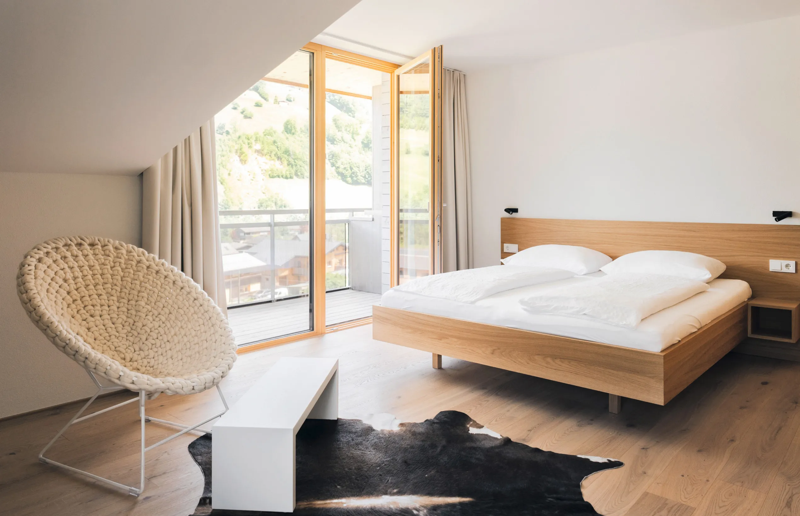 Helles, modernes Schlafzimmer im Aparthotel Hubertus in Mellau mit gemütlichem Doppelbett, Designer-Sessel und direktem Zugang zum Balkon mit beeindruckendem Bergblick über den Bregenzerwald.