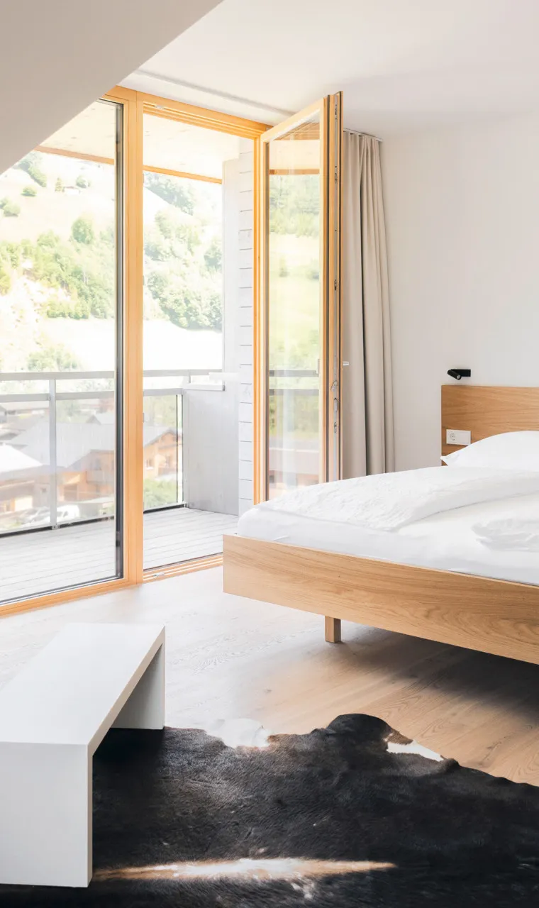 Helles, modernes Schlafzimmer im Aparthotel Hubertus in Mellau mit gemütlichem Doppelbett, Designer-Sessel und direktem Zugang zum Balkon mit beeindruckendem Bergblick über den Bregenzerwald.