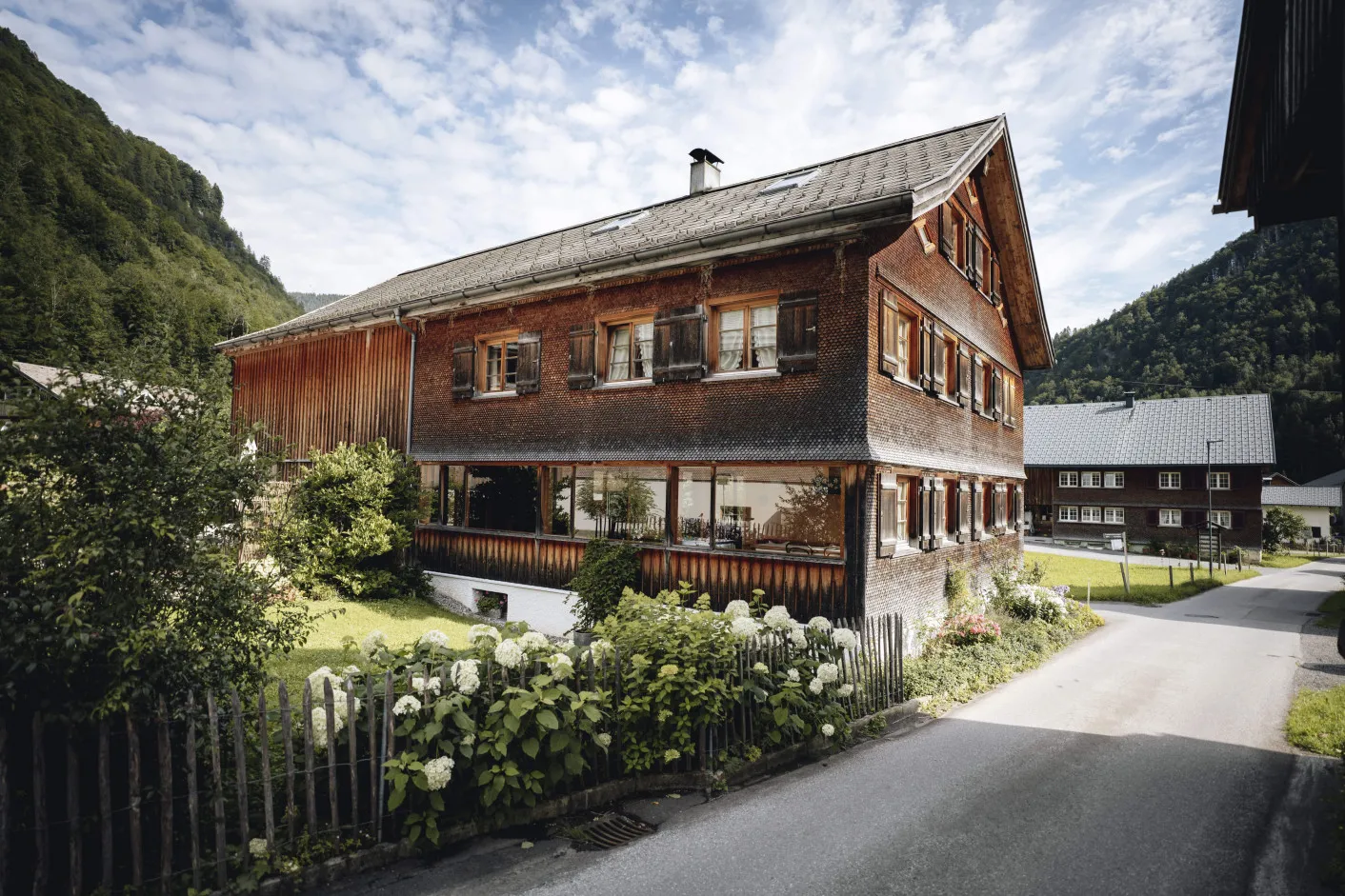 Traditionelles Holzhaus mit charakteristischer Architektur und Blumen im Vordergrund, umgeben von der malerischen Berglandschaft von Mellau, das die charmante Umgebung und die lokale Bauweise nahe dem Aparthotel Hubertus widerspiegelt.