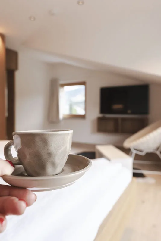 Hand hält eine Kaffeetasse in einem hellen Apartment des Aparthotel Hubertus in Mellau, mit Blick auf den Wohnbereich mit Fernseher und Fenster mit Bergblick – symbolisiert gemütliche Morgenstunden und Entspannung im Bregenzerwald.