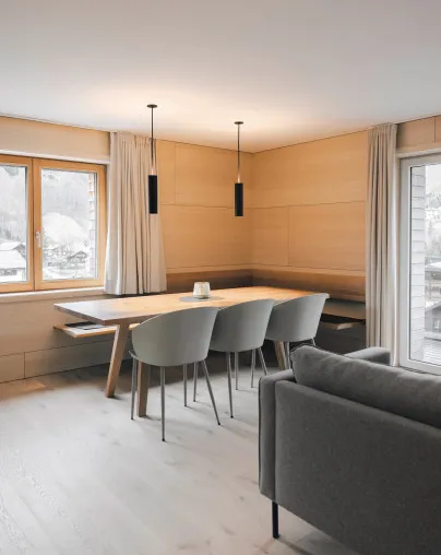 Gemütlicher Essbereich in einem Apartment des Aparthotel Hubertus in Mellau – mit einem Holztisch, integrierter Bank, modernen Stühlen und zwei Pendelleuchten, daneben ein großes Fenster mit malerischem Blick auf die verschneite Bregenzerwald-Landschaft.