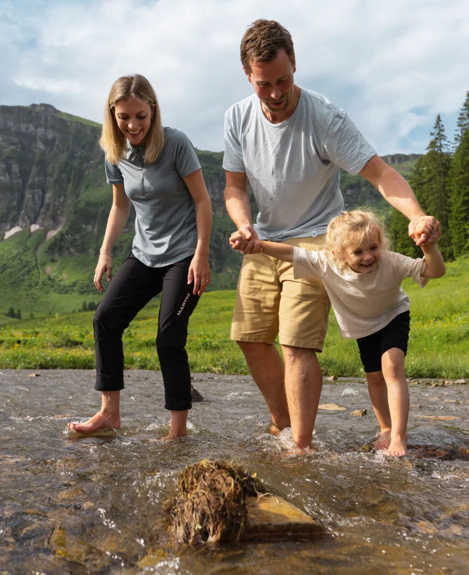 Eine glückliche Familie, bestehend aus Eltern und Kind, watet barfuß lachend durch einen erfrischenden Bergbach in den grünen Alpen des Bregenzerwaldes – ein ideales Sommererlebnis für Familien, die ihren Urlaub im Aparthotel Hubertus in Mellau verbringen.