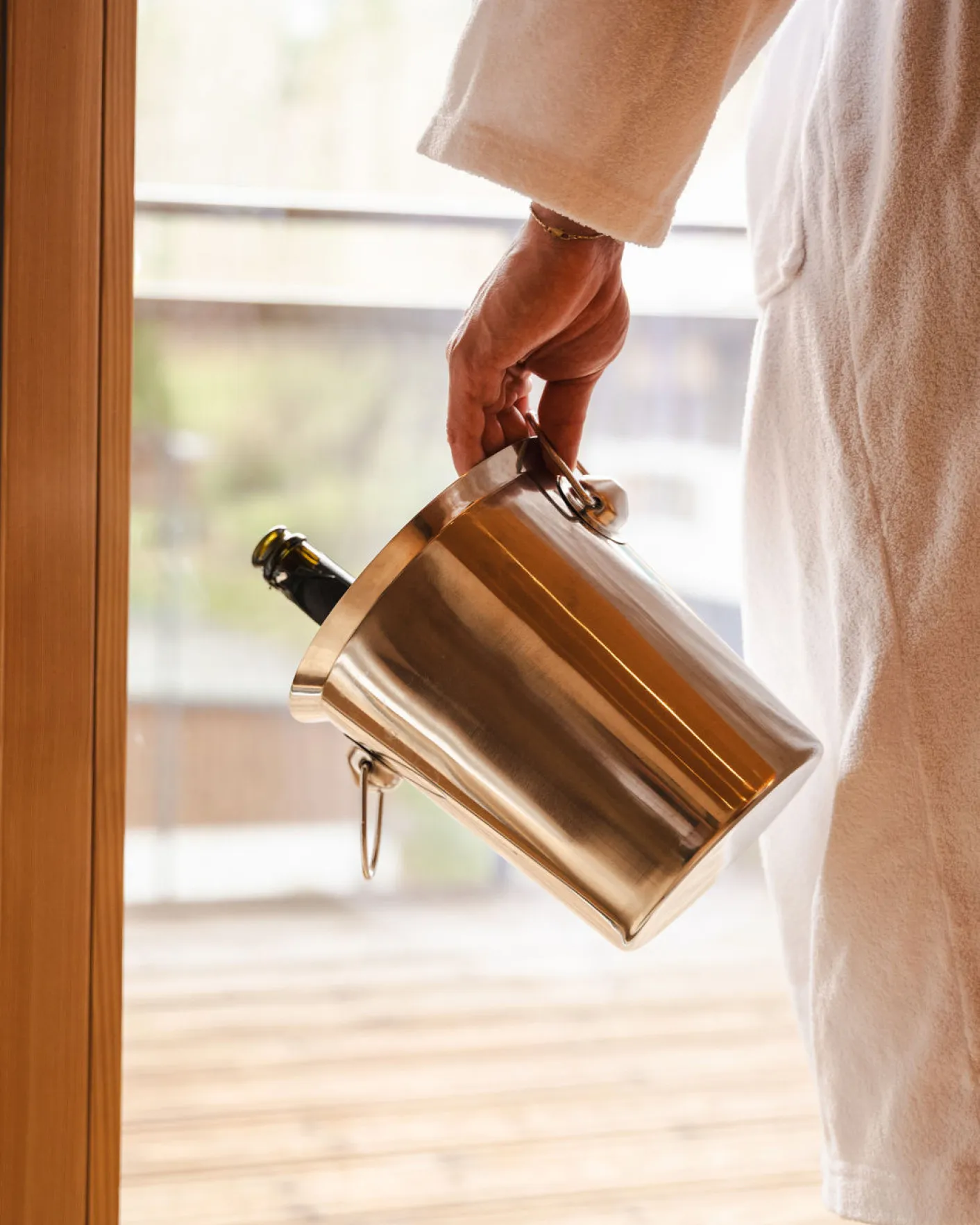Ein Gast im Bademantel des Aparthotel Hubertus in Mellau hält einen Sektkühler mit einer Flasche, bereit für einen luxuriösen Genussmoment auf dem Balkon mit Blick auf die Natur des Bregenzerwaldes.