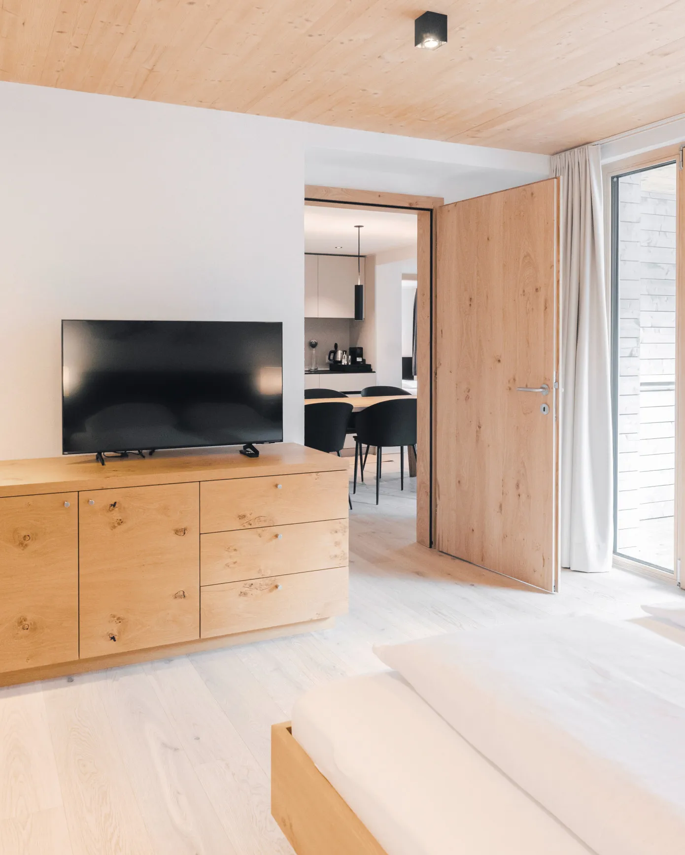 Helles Schlafzimmer in einem Apartment des Aparthotel Hubertus in Mellau mit modernem Holzschrank, Flachbildfernseher und Blick in den offenen Wohnbereich mit Essplatz und Küche – ideal für Gäste, die Komfort und Privatsphäre im Bregenzerwald schätzen.