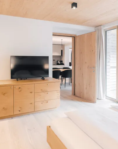 Helles Schlafzimmer in einem Apartment des Aparthotel Hubertus in Mellau mit modernem Holzschrank, Flachbildfernseher und Blick in den offenen Wohnbereich mit Essplatz und Küche – ideal für Gäste, die Komfort und Privatsphäre im Bregenzerwald schätzen.