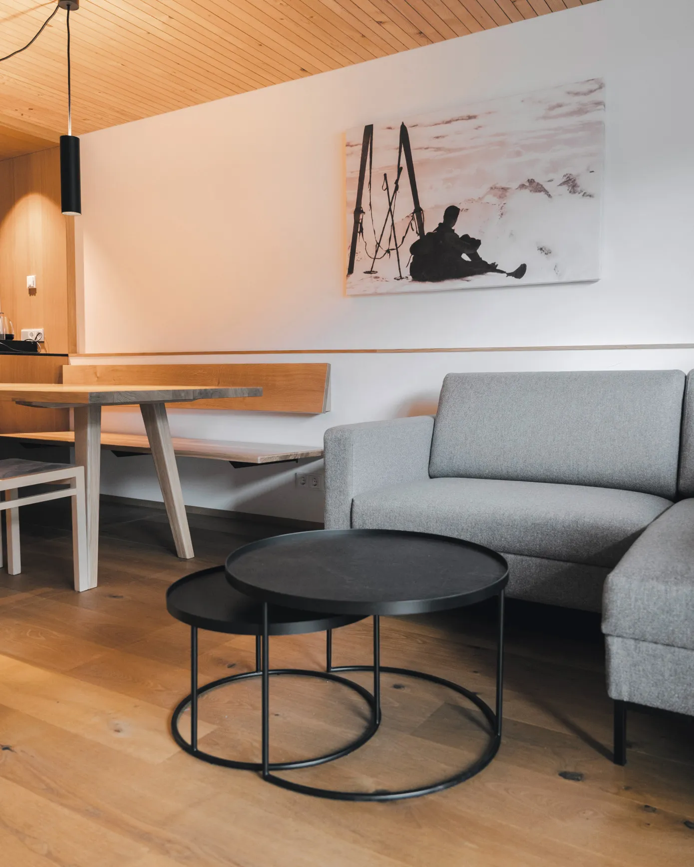 Offener Wohn- und Essbereich in einem Apartment des Aparthotel Hubertus in Mellau – mit gemütlichem Sofa, modernen Couchtischen, einem Wandbild mit Skimotiv und einem Essplatz mit Holztisch und Bank, perfekt für einen entspannten Winterurlaub im Bregenzerwald.