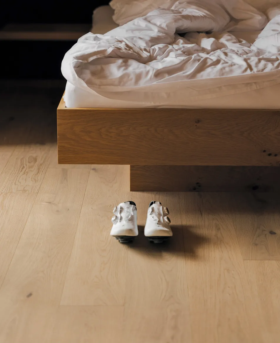 Ein gemütliches Doppelbett im Aparthotel Hubertus in Mellau, mit bereitstehenden Radschuhen auf dem Holzparkettboden – symbolisiert den idealen Komfort nach einem aktiven Radausflug im Herzen des Bregenzerwaldes.