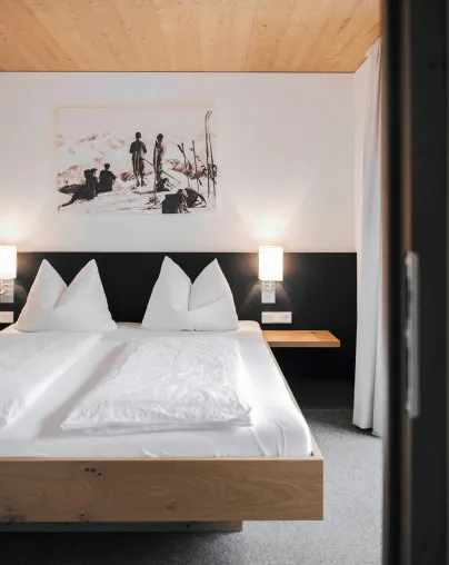 Stilvolles Schlafzimmer im Aparthotel Hubertus in Mellau mit modernem Doppelbett, Holzbalkendecke und einem Wandbild von Skifahrern – eine gelungene Kombination aus alpinem Charme und zeitgenössischem Design für Ihren Urlaub im Bregenzerwald.