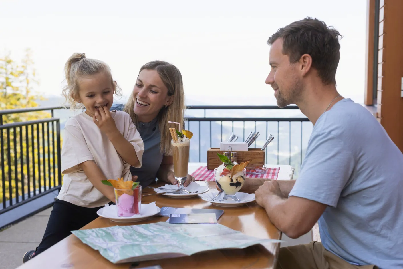 Eine glückliche Familie genießt Eis und Getränke auf einer Sonnenterrasse, mit einem Wanderkarte auf dem Tisch und Blick auf die umliegenden Alpen – perfekt für einen Familienurlaub und Outdoor-Aktivitäten im Bregenzerwald.