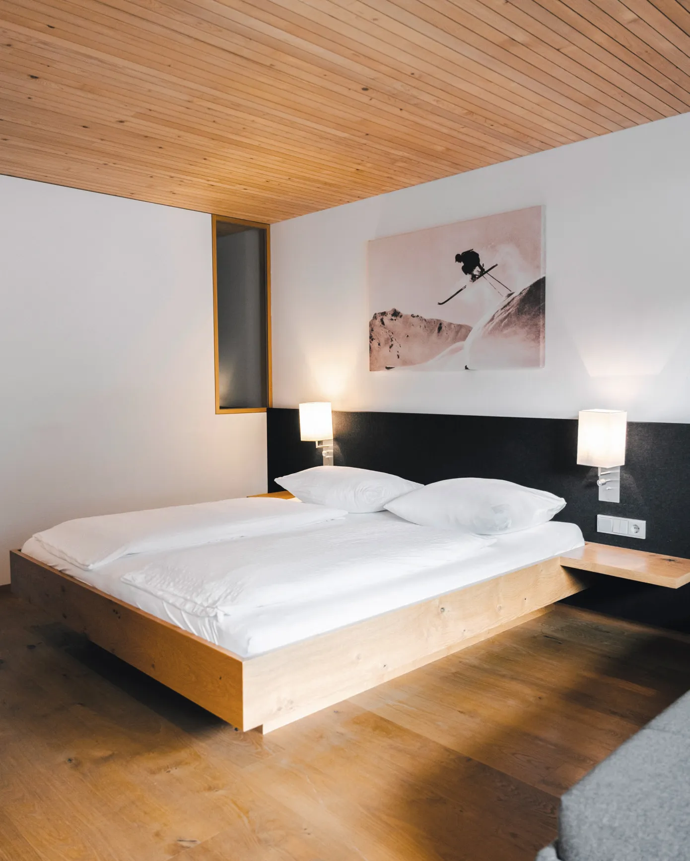 Stilvolles Schlafzimmer im Aparthotel Hubertus in Mellau mit modernem Doppelbett, Holzbalkendecke und einem beeindruckenden Wandbild eines Skifahrers – eine Hommage an die Vorarlberger Alpen und perfekt für aktive Urlauber im Bregenzerwald.