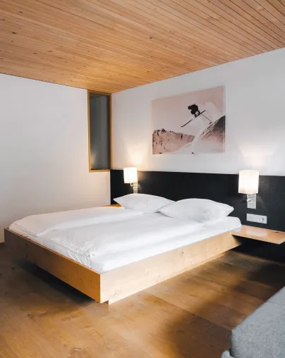 Stilvolles Schlafzimmer im Aparthotel Hubertus in Mellau mit modernem Doppelbett, Holzbalkendecke und einem beeindruckenden Wandbild eines Skifahrers – eine Hommage an die Vorarlberger Alpen und perfekt für aktive Urlauber im Bregenzerwald.