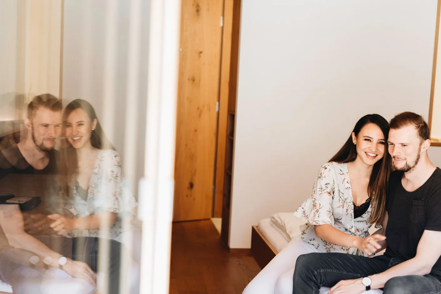Ein glückliches Paar genießt entspannte Momente in einem modernen Apartment des Aparthotel Hubertus in Mellau, gespiegelt in einer Glasfläche – perfekt, um romantische Urlaube und Komfort zu zweit im Bregenzerwald zu bewerben.