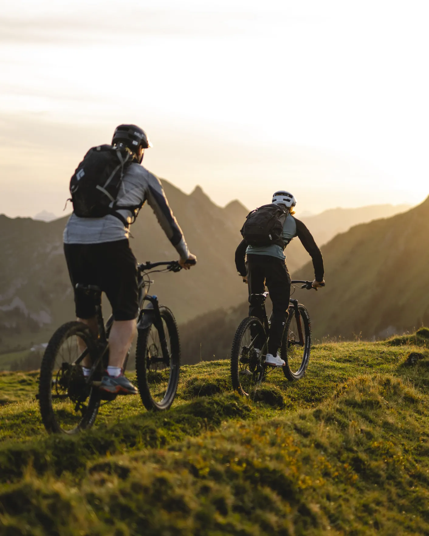 Zwei Mountainbiker auf einem Bergpfad im Bregenzerwald bei Sonnenuntergang, mit der beeindruckenden Kulisse der Alpen – perfekt, um die vielfältigen Radfahrmöglichkeiten rund um das Aparthotel Hubertus in Mellau zu präsentieren.