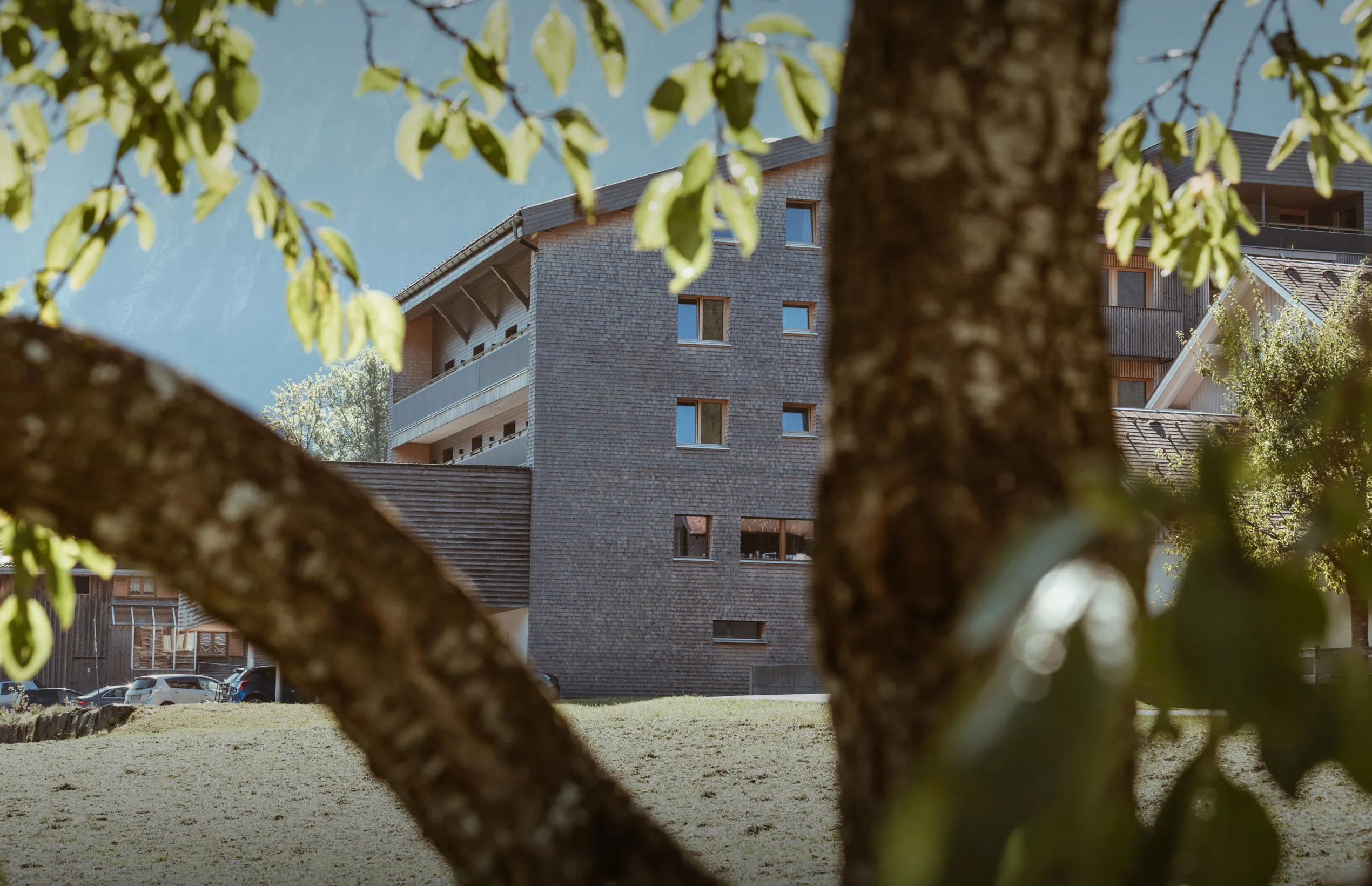 Die moderne Außenansicht des Aparthotel Hubertus in Mellau im Sommer, umgeben von grünen Wiesen und Bäumen, mit seiner charakteristischen Holzschindelfassade – perfekt für Ihren erholsamen Urlaub im Bregenzerwald.