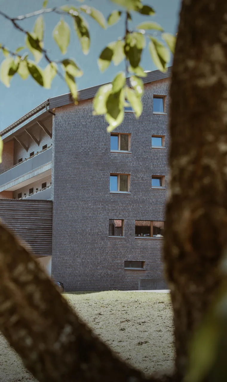 Die moderne Außenansicht des Aparthotel Hubertus in Mellau im Sommer, umgeben von grünen Wiesen und Bäumen, mit seiner charakteristischen Holzschindelfassade – perfekt für Ihren erholsamen Urlaub im Bregenzerwald.