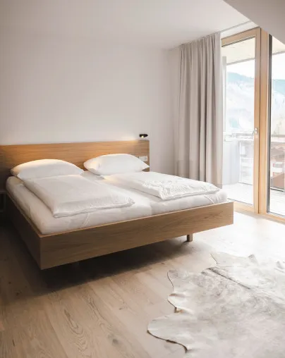 Schlafzimmer in einem Apartment des Aparthotel Hubertus in Mellau mit modernem Doppelbett, Holzboden, Fellteppich und Zugang zum Balkon mit Bergblick – einladend und komfortabel für erholsamen Urlaub im Bregenzerwald.