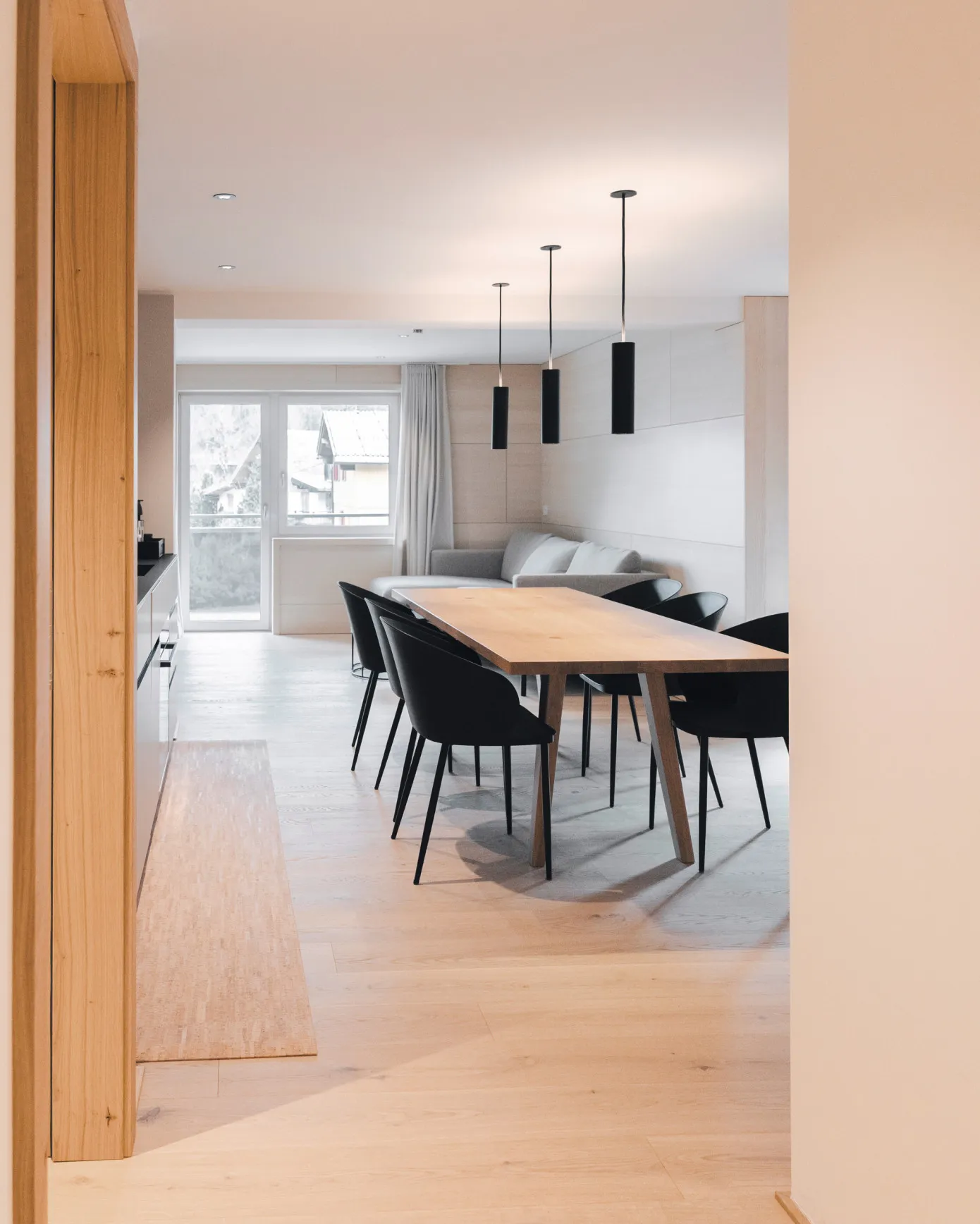 Großzügiger offener Wohn-, Ess- und Küchenbereich in einem Apartment des Aparthotel Hubertus in Mellau – mit modernem Esstisch, schwarzen Stühlen, stylischen Pendelleuchten und einem bequemen Sofa, ideal für einen erholsamen Aufenthalt im Bregenzerwald.
