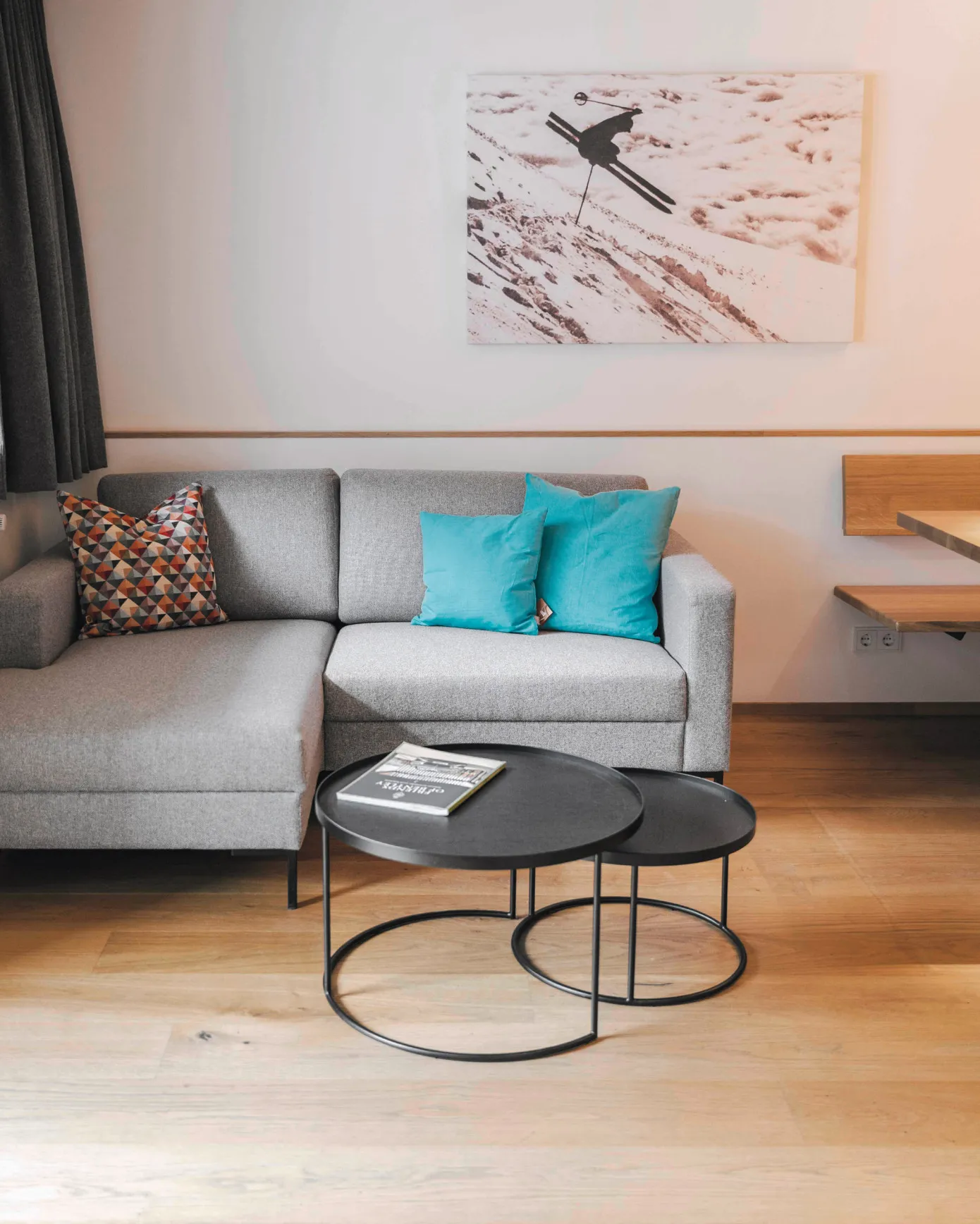 Gemütliches Wohnzimmer in einem Apartment des Aparthotel Hubertus in Mellau mit grauem Sofa, blauen Kissen und einem Wandbild von Skispuren im Schnee – der ideale Ort zum Entspannen nach einem Tag auf den Pisten des Bregenzerwaldes.