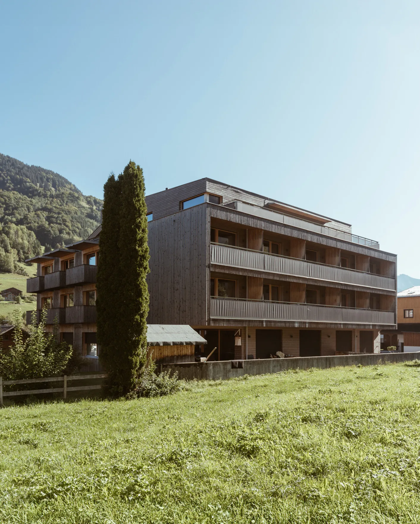 Die moderne Außenansicht des Aparthotel Hubertus in Mellau im Sommer, umgeben von grünen Wiesen und Bäumen, mit seiner charakteristischen Holzschindelfassade – perfekt für Ihren erholsamen Urlaub im Bregenzerwald.